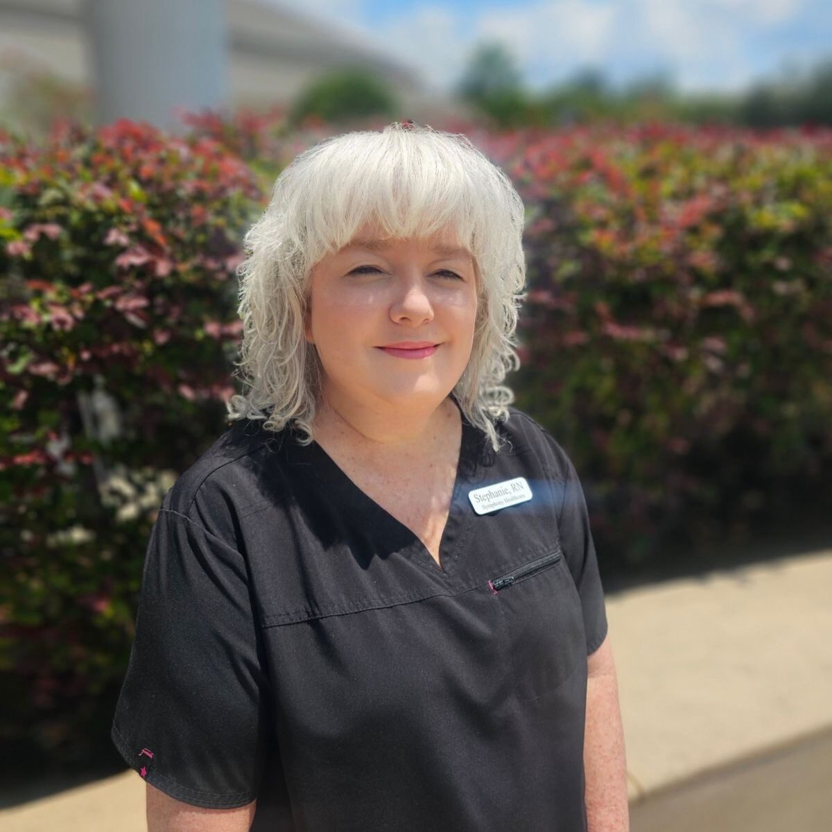 Stephanie A woman with white hair standing in front of bushes.